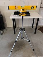 Full view showing yellow rotating laser level mounted on adjustable tripod with spirit level bar on top and carrying case on table behind