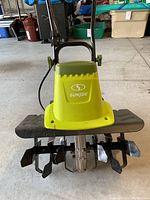 Front view of Sun Joe tiller showing green and black body, Sun Joe branding, and multiple metal tilling blades.