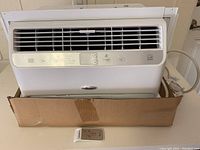 Whirlpool window air conditioner in a cardboard box showing unit front with vents, control panel buttons, and remote control in front.