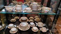 Upper and lower glass shelves displaying full set of Famille Rose porcelain pieces including stacked plates, bowls, cups, saucers, teapot, sugar bowl and creamer.