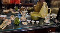 Various vintage Asian ceramics and kitchenware items displayed, including two floral teapots, ceramic wonton spoons, sake cups, small bowls, a green crackle glass pitcher, cream and olive-green pitchers, and an enameled cast iron baking dish.