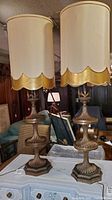 Pair of vintage tiered brass fountain-style lamps with beige and gold scalloped lampshades on a white painted chest in room setting.