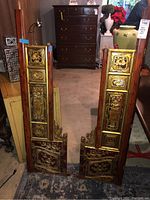 Two ornate wooden bed parts with detailed floral carvings and gold decoration, placed upright on floor and leaning against furniture