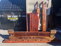 Five wooden Chinese wedding bed pieces photographed outdoors, showing painted and carved designs in red and gold colors. Pieces are laid out against a dumpster as background.