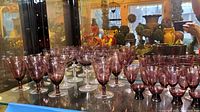 Wide view showing multiple amethyst colored crystal stemware with clear stems arranged on glass shelf.