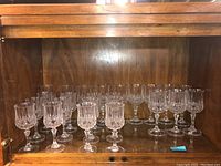 Set of 24 crystal stemware glasses arranged on a wooden shelf inside a cabinet, showing multiple glasses of similar size with intricate cut glass designs.