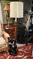 Wooden floor lamp with adjustable height and cream fabric lampshade beside black lacquered wooden kissing couple sculpture on a red patterned carpet.