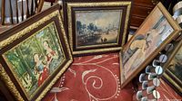 Three framed prints shown leaning against furniture on a red patterned carpet, two larger prints feature outdoor scenes and one smaller print with a nude female figure.