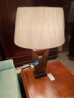 Full view of lamp with beige fabric shade on metal geometric base, placed on wooden surface near blue upholstered chair.