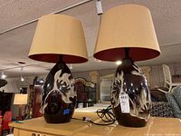 Two ceramic base lamps with beige shades on a wooden surface in an indoor environment. Both lamps have an oval shape with brown and white abstract organic design bases and visible sticker residues. Beige drum shades with red inner lining.