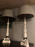Pair of white table lamps with gray drum shades placed side by side on a black surface against a gray wall. Lamps have detailed carved bases with claw foot design. Tags attached to the cords.