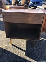 Front view of a rectangular mid-century modern side table with a drawer and open shelf, wood finish, and angled legs.