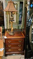 Front and side view of a wooden Dixie brand nightstand with three drawers, showing wood grain finish and brass hardware, alongside a lamp with a giraffe under a palm tree on the base and a tapered fabric lampshade.