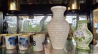 Photo showing 8 artisan pottery cups in front of 2 decorative vases on glass shelving.