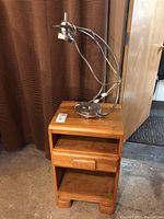 Side view of blond wood side table with drawer and open shelves, and desk lamp on top.