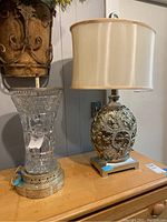 Pair of two table lamps on wooden surface. Left lamp has clear crystal base with gold metal bottom, right lamp has ornate gold base with beige fabric shade.