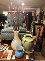 Wide shot of three vases in room setting with furniture in background. Cream-colored large vase, yellow ceramic vase, and blue ceramic pitcher.