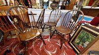 Set of 4 brown wooden Windsor chairs with curved spindled backs and contoured wooden seats, showing scratches and patina. Positioned on a red patterned carpet with other furniture items around.