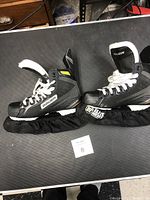 Pair of black Bauer ice hockey skates placed on a textured surface, showing both skates and blade covers.