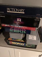 Stacked board games including Pictionary, Rummikub, Scruples, Triopoly, and others with gray plastic cases