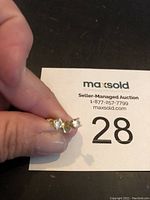 Front view of the pair of three-stone diamond stud earrings held in hand against a black surface with MaxSold card