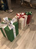 Three decorated presents in green, white, and red glitter with ribbons and metallic ball bows, set indoors on wood floor.