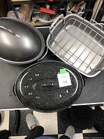 Three roasting pans shown: one black oval pan with lid, one Kitchen Aid pan with rack, one black classic pan with raised bottom and lid. Pans are on a black surface, surrounding items are visible.