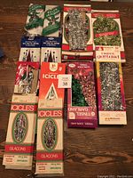 Photo showing the full assortment of tinsel and garland packages laid out on a wood floor, with visible vintage packaging from Yulecraft and Woolworths, including colorful and silver icicles.