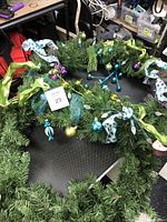 Three artificial wreaths on a table: one green with blue/green ornaments and ribbons, one colorful with pink, blue and purple decorations, one plain green wreath