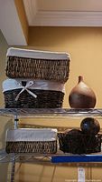 Three rectangular woven baskets with white cloth liners stacked on a metal shelf, with a large textured brown vase on the right side of the top shelf. Below, a heart shaped basket with decorative spheres and one rectangular basket are visible.