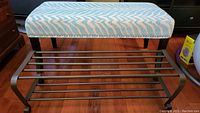 Overall view of upholstered bench with blue and white fabric and stud detailing beside black metal shoe rack on wooden floor.