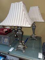Pair of table lamps with metal bases and cream striped fabric shades on a glass surface. One lamp's base is fully visible showing decorative detailing and claw-foot style legs.