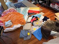 Photo showing an assortment of folded and unfolded linens including duvets, sheets, throws, pillow cases, and a small rug arranged on the floor and furniture.