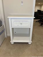 Front view of the white wooden nightstand showing one drawer with knob and open shelf below.
