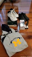 Lot items on wood floor showing golf shoes on box, black slip-on moc shoes, bobblehead, and folded Michigan football hoodie.