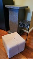 Photo shows the full view of all three items: the large stone/concrete display pedestal stand in gray speckled finish, the black metal ornamental plant stand with a rectangular top, and the beige fabric ottoman stool with a buttoned top. Positioned on hardwood floor near wall.