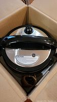 Top view of Power Cooker XL Deluxe black and stainless steel lid with pressure valve and caution label inside cardboard box