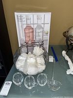 Glass punch bowl with 3 glass cups in front, punch bowl filled with paper towels for protection, glass punch serving spoon to the right, boxed Fiddle Fern cold drink dispensers in the background.