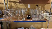 Metal wire shelf with assorted stemware and bar glassware arranged including wine, martini, Crown Royal cocktail glasses, mugs, and a cocktail pitcher with a lid.