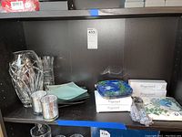 Shelf display of all items including crystal vases, candle holders, stacked green serving plates, eight boxed Fragonard glass plates, spoons, and napkins.