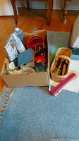 Photo of open box showing art supplies including paint tubes, pencils, red paint palette, and wooden pencil box on carpeted floor.
