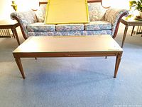Full view of light brown rectangular Mid Century Modern wood coffee table with tapered legs in front of sofa.