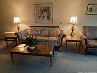 Wide shot showing two wood end tables on either side of a floral-patterned couch with lamps on each table, along with a larger wood coffee table in front. Shows table design, size, and color relative to furniture.