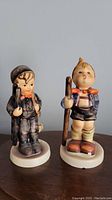 Two Hummel figurines side by side on a wooden surface: "Chimney Sweep" and "Little Hiker" with visible chips on base.