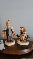 Two Hummel figurines, a boy holding boots and a girl sitting on a fence with basket of apples.