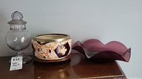 Lot showing antique Imari bowl with silver rim, vintage glass pedestal candy bowl with lid, and purple art glass display bowl on wooden surface