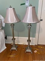 Pair of tall lamps with light beige fabric shades on a wooden floor in front of a green wall.