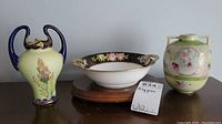Wide view of lot showing three ceramic pieces: a small blue-handled yellow vase, a floral dark bowl, and a decorated urn vase with floral ovals and green-pink background.