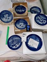 Seven Royal Copenhagen blue and white collector plates laid out on a table, including packaging foam and cardboard boxes.