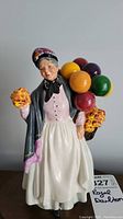 Front view of the Royal Doulton Biddy Pennyfarthing figurine showing detailed facial expression, bonnet, shawl, pink blouse, white skirt with basket of colorful balls and flowers.